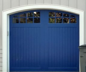 Blue wooden garage door with two arched windows on white vinyl garage